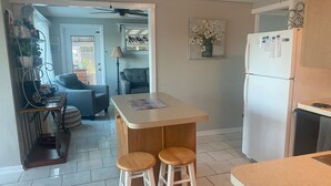 Cocina compacta con refrigerador, una barra pequeña con taburetes, ventilador de techo y comedor con sofá y lámpara.