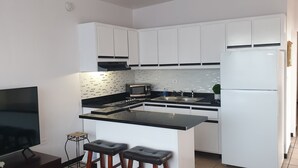 Une cuisine de taille réduite avec des meubles blancs, un plan de travail noir, un four à micro-ondes, un évier et un réfrigérateur. On y trouve également deux tabourets et un téléviseur.