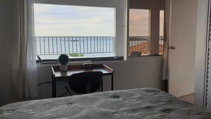 Une chambre à coucher avec un lit, un bureau et une chaise, une petite plante en pot et une fenêtre donnant sur la mer.