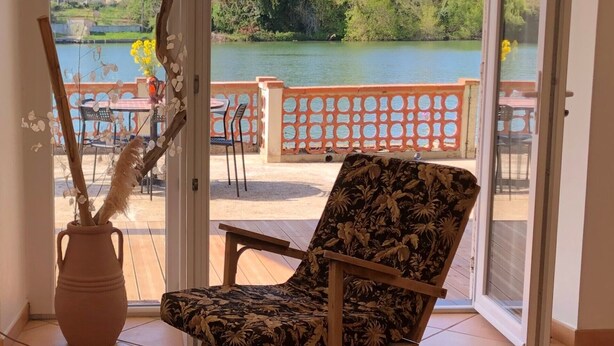 A room with a view of a lake, a chair, and a vase with dried flowers.