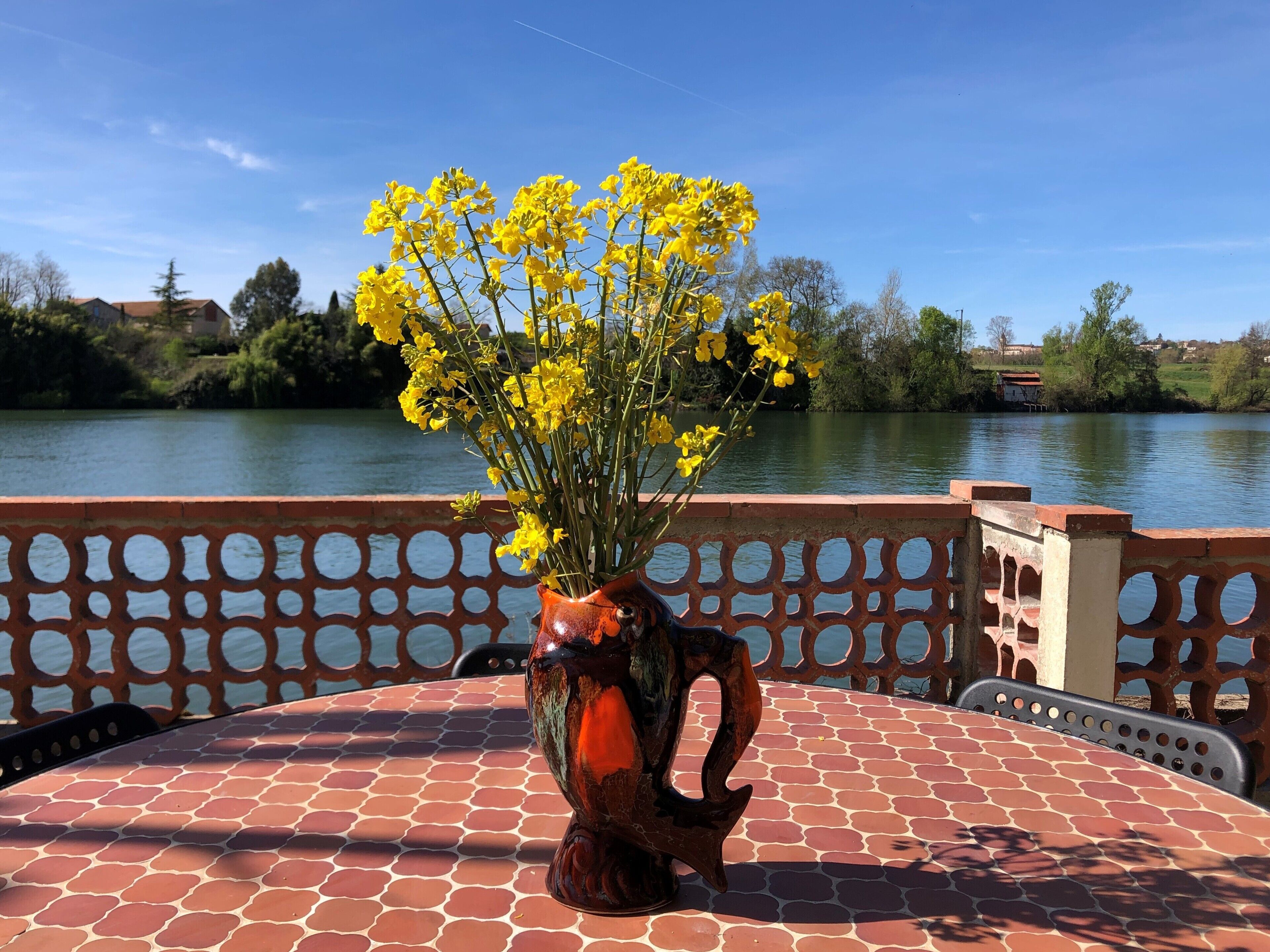 A table with a vase of yellow flowers, a patterned tablecloth, and a view of a body of water with a fence in the foreground.
