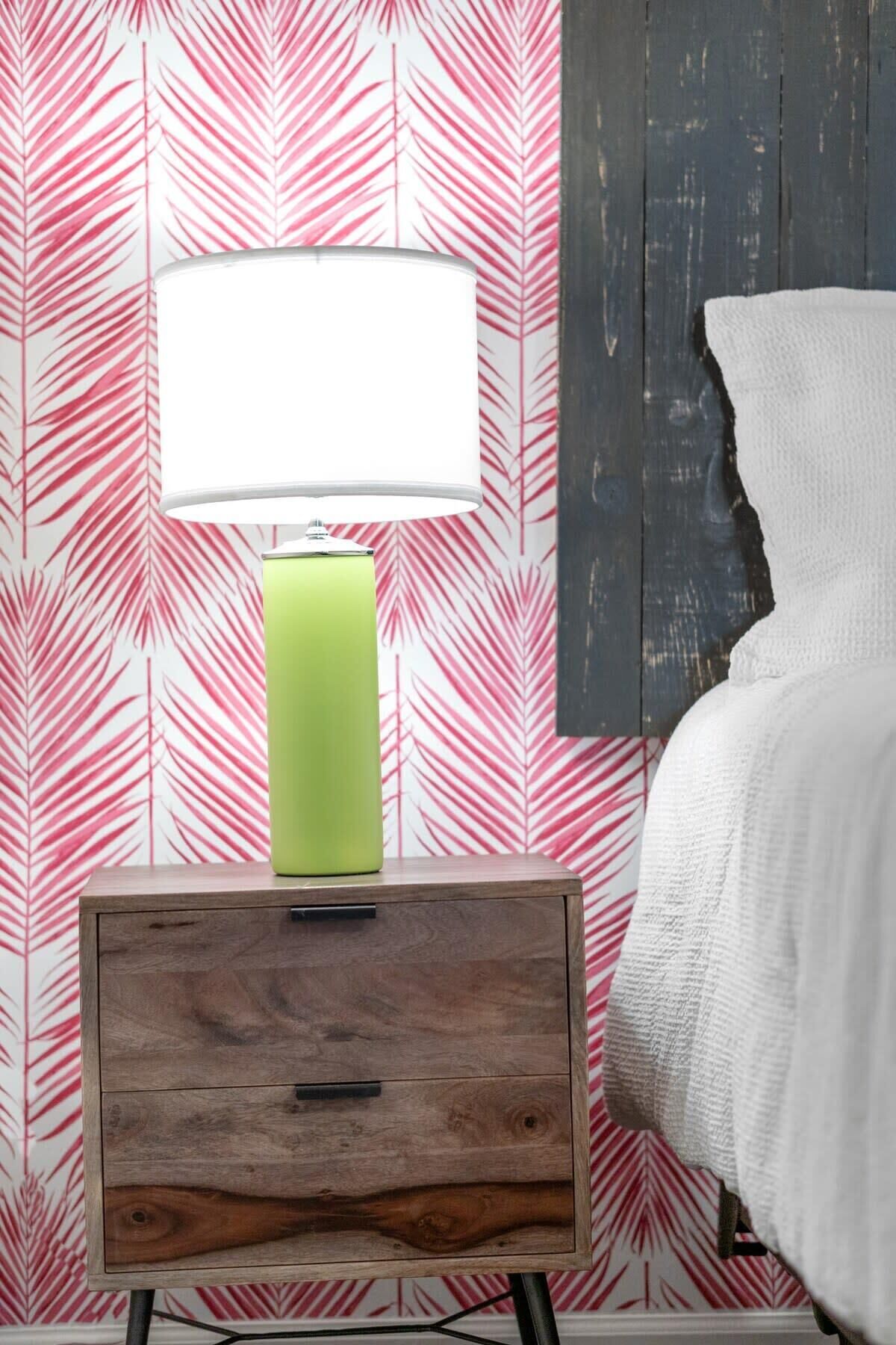 Una cama con una manta blanca, una lámpara verde sobre una mesita de noche de madera, y una pared con un diseño de hojas rosas.