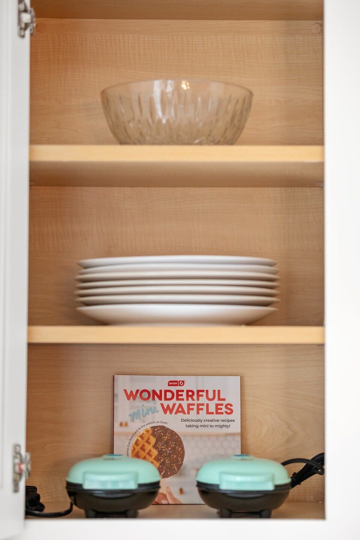 Een keukenkastje met een glazen schaal, gestapelde borden en een kookboek met de titel 'WONDERFUL mini WAFFLES'.