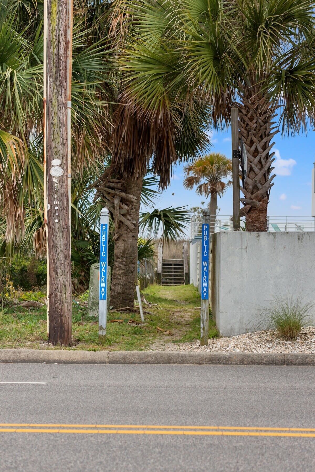 A street sign indicating a public walkway adjacent to a road, with palm trees and a concrete wall in the background.