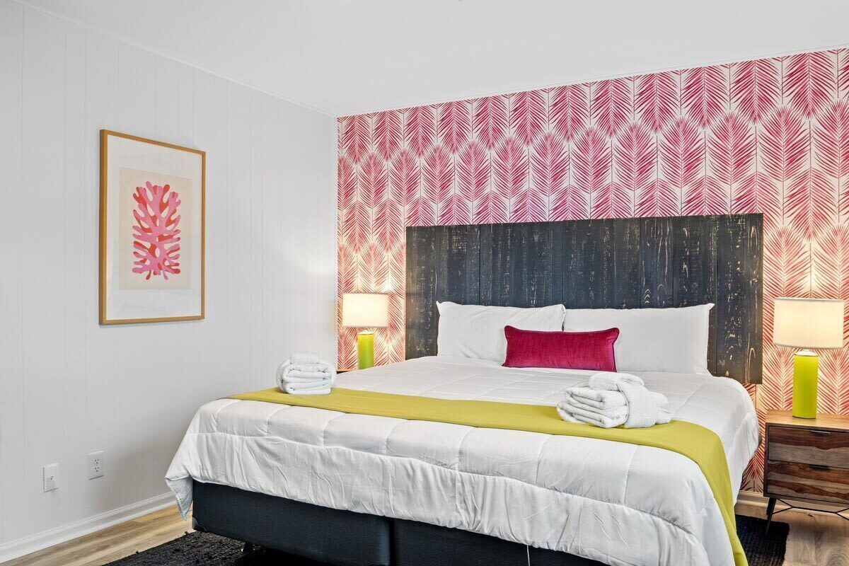 A hotel room with a large bed, white bedding, a red pillow, and a patterned wallpaper. There are two bedside tables with lamps.