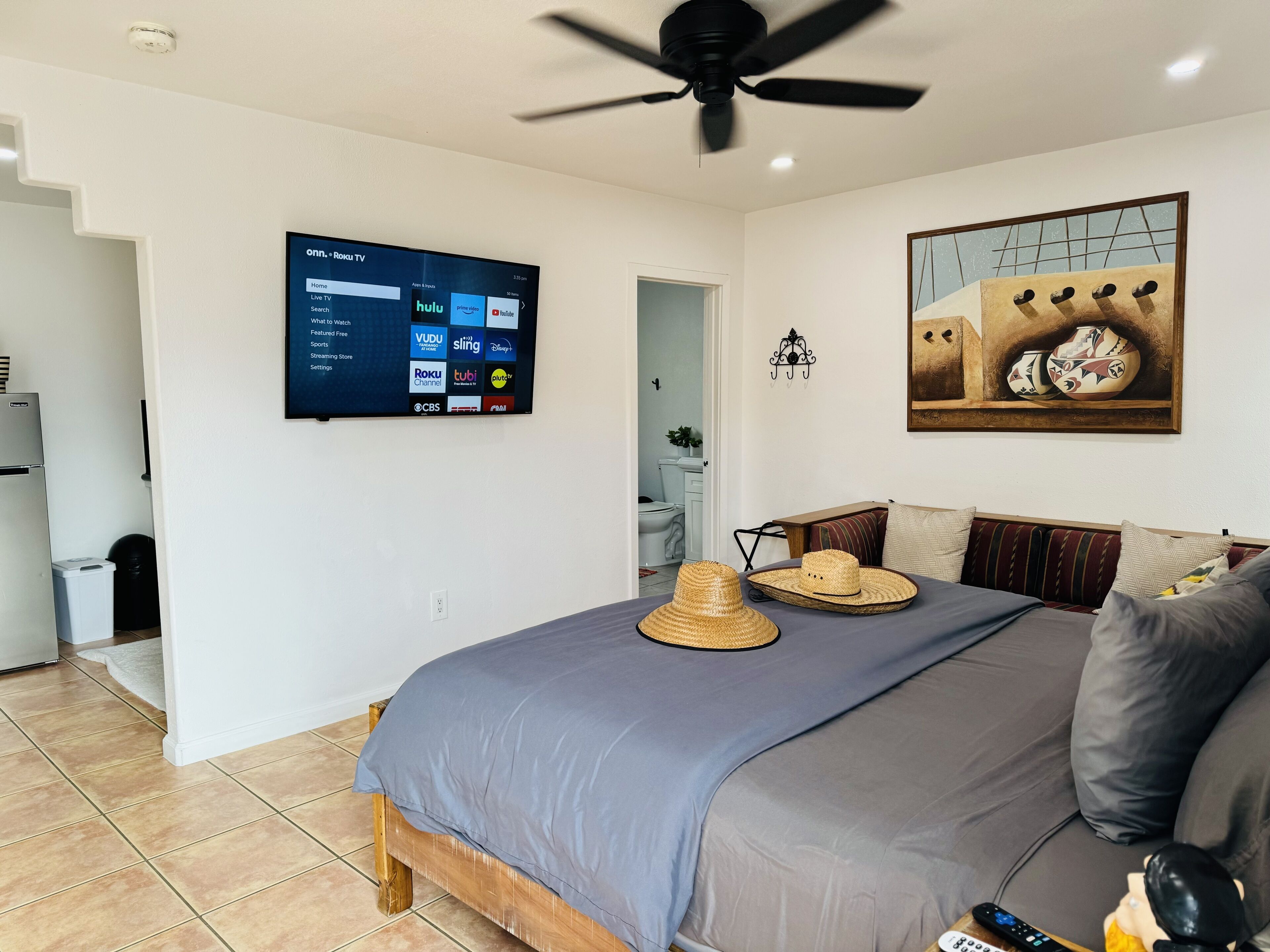 A bedroom with a bed, a ceiling fan, a mounted TV displaying streaming services, a painting, and a remote control on the bed.