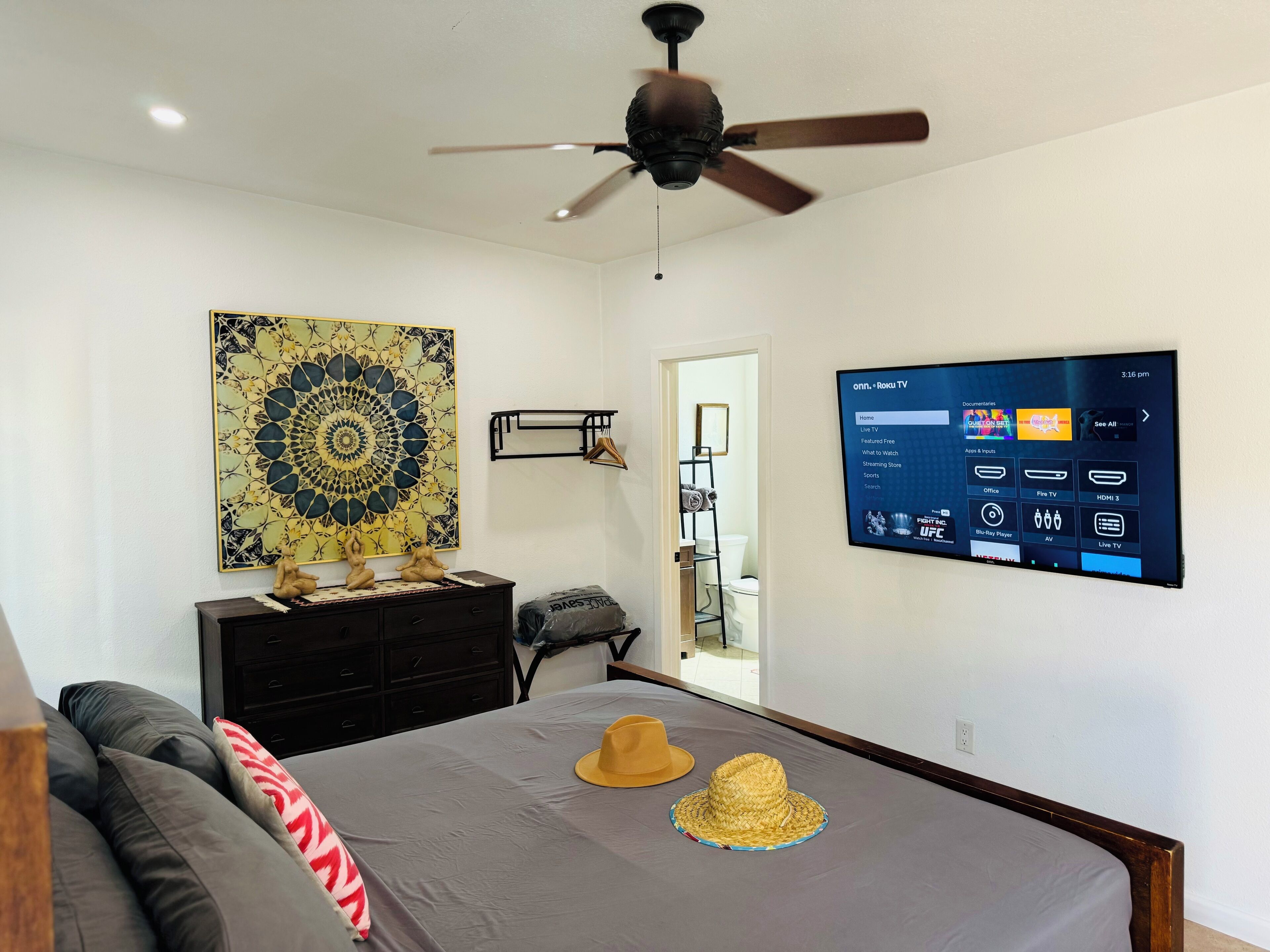 A bedroom with a bed, a ceiling fan, a wall-mounted TV, a decorative wall piece, and a dresser.