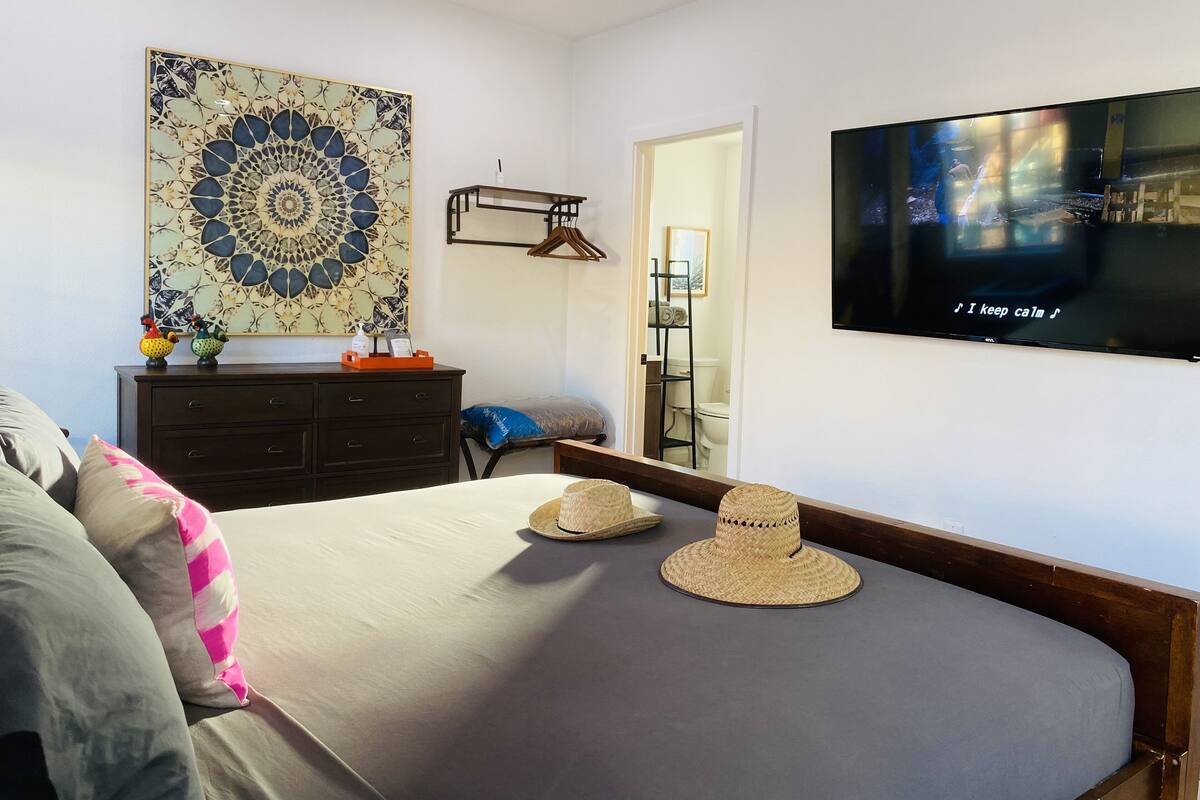 Une chambre à coucher comprenant un lit, un téléviseur, une commode, un élément décoratif mural, et une salle de bain visible à travers une porte ouverte.