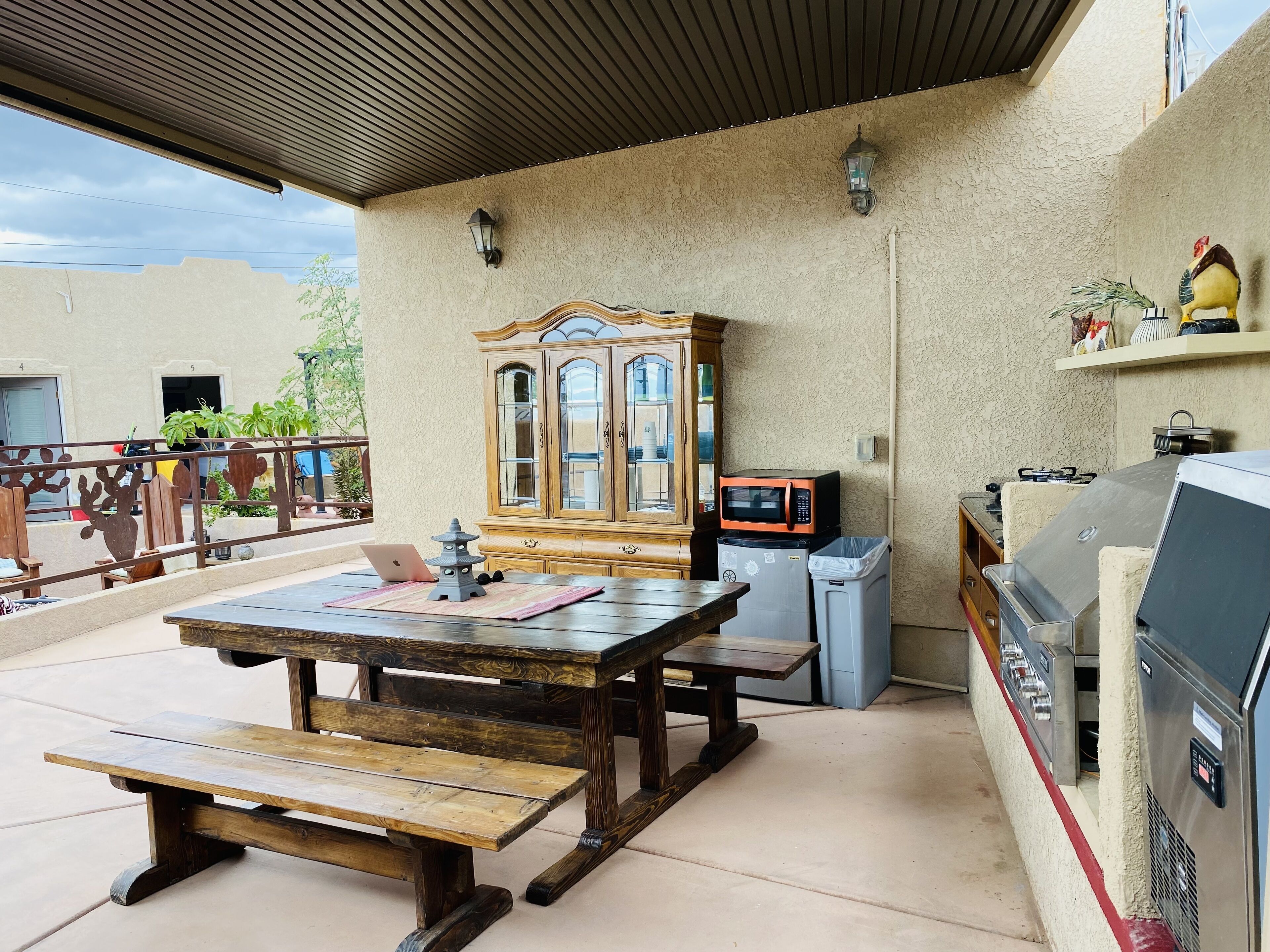 An outdoor patio with a wooden table and benches, a cabinet, a microwave, a trash bin, and a grill.