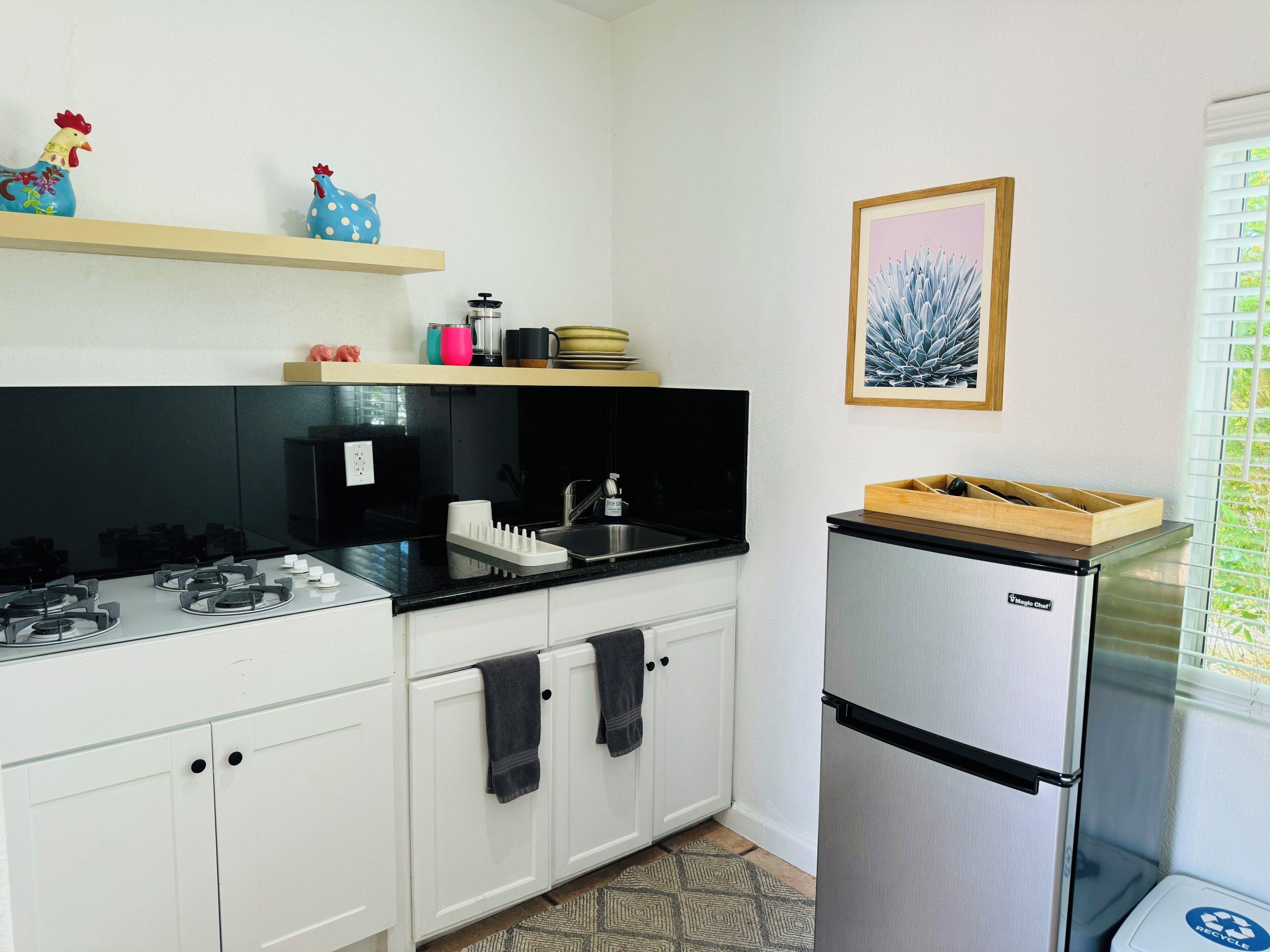 A modern kitchen with white cabinets, a black countertop, a stainless steel refrigerator, and a window with blinds.