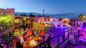 Uma área de piscina ao ar livre com espreguiçadeiras e uma lareira, rodeada por edifícios com iluminação quente.