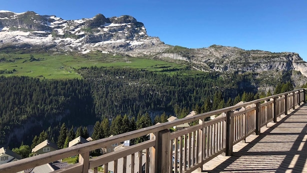A wooden deck with a view of a mountainous landscape, including a forested valley and rocky peaks.