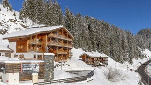 A large wooden lodge with multiple balconies and a stone entrance, surrounded by snow-covered trees and a clear sky.