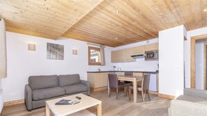 A compact living space with a wooden ceiling, a grey sofa, a dining table with chairs, and a kitchen area with a microwave.