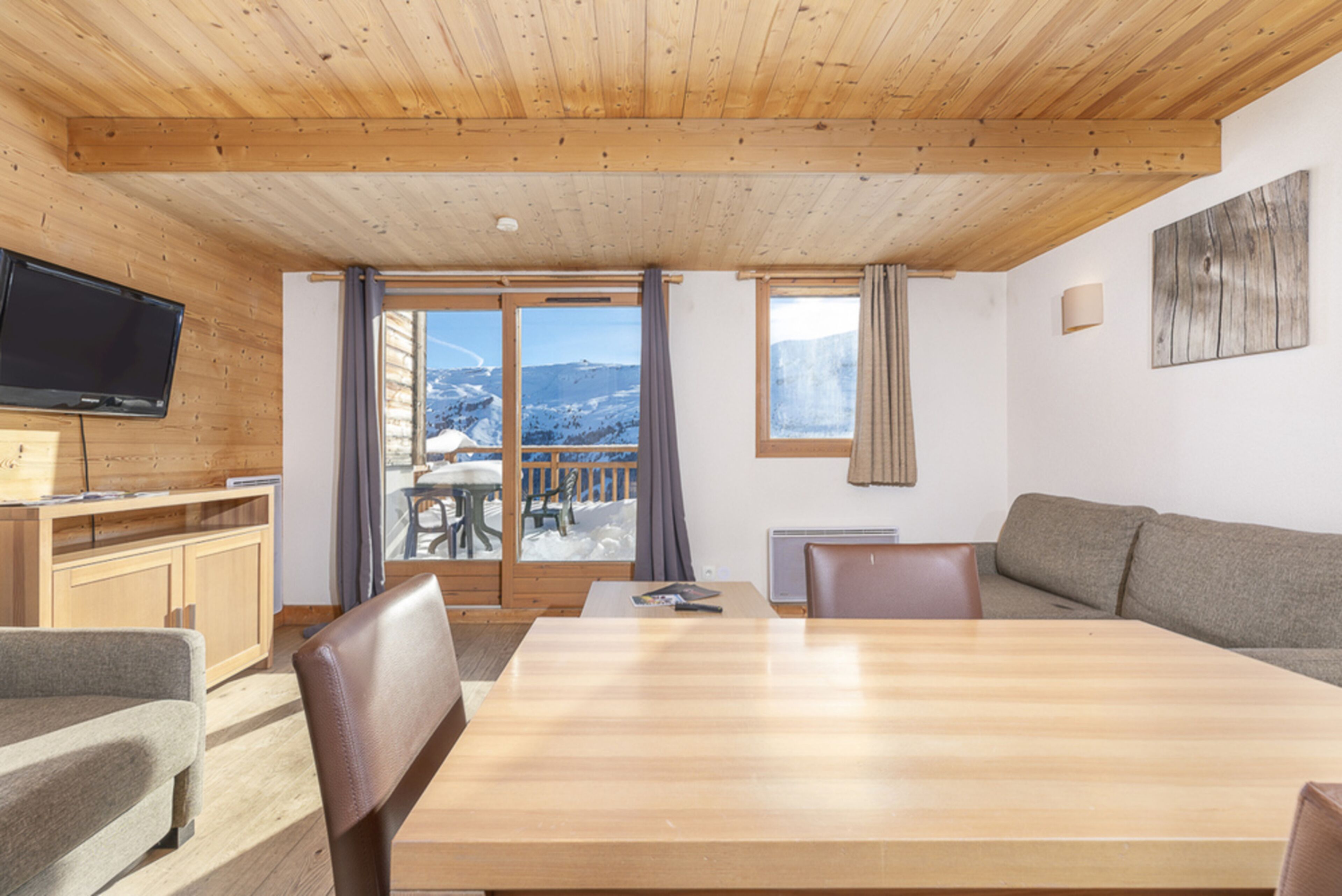 A wooden-paneled room with a dining table, chairs, a sofa, a television, and a view of snowy mountains through large windows.