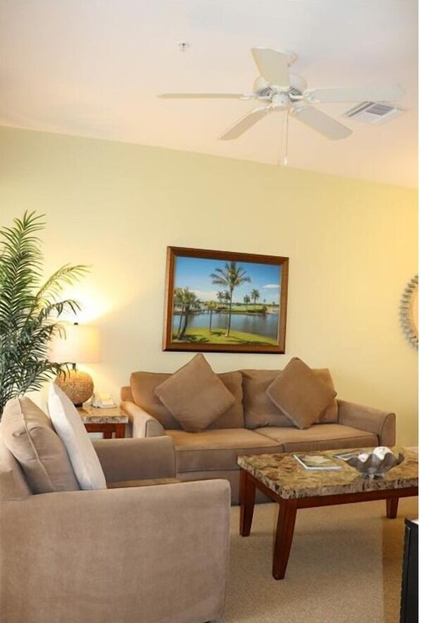 A living room with a beige sofa, a coffee table, a ceiling fan, and a framed picture of a beach scene. - Lely Resort 7965 Mahogony Rn Lane # 325- 2 Bedroom Golf Course View (Naples)