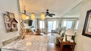 A kitchen with a granite countertop, wooden cabinets, and pendant lights. There is a dining area with a table and chairs, and a living room with a couch and a lamp.