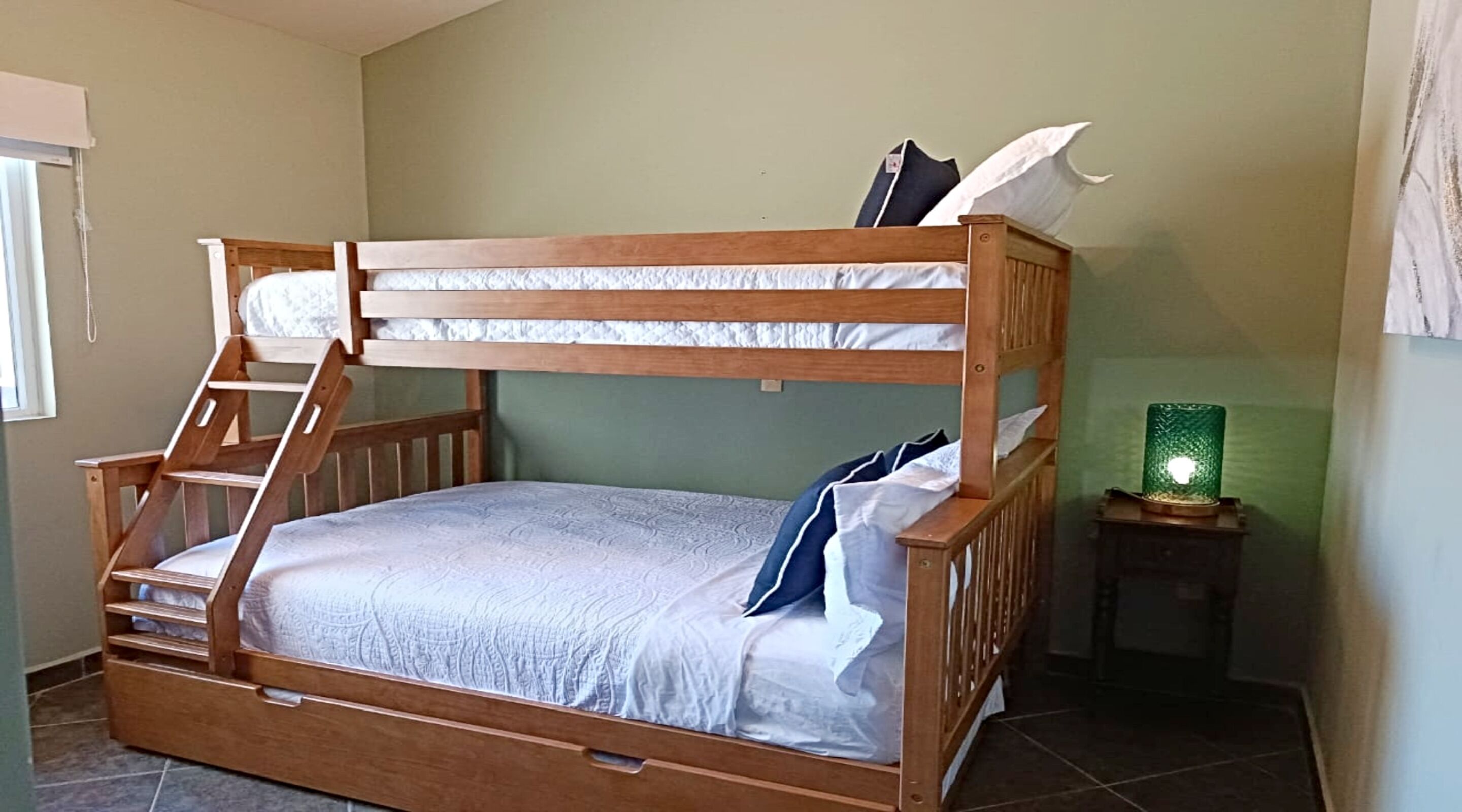 Een stapelbed met een ladder, een klein nachtkastje met een groene lamp en een raam met zonwering.