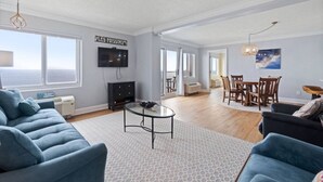 Een ruime woonkamer met een groot raam, een flatscreen televisie, een eethoek met houten meubels en een plafond met een decoratieve lamp.