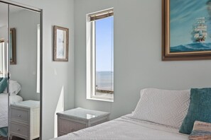 A bedroom with a bed, a nightstand, a dresser, a large mirror, and a window with a view of the sea.