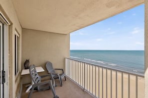 A balcony with a table and two chairs, overlooking a beach with the ocean.