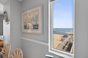 A room with a large window overlooking a beach, a framed artwork on the wall, and a chair.