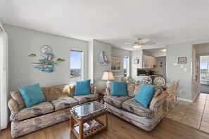 A living room with two patterned sofas, a glass coffee table, and a view of the kitchen and outdoor area.