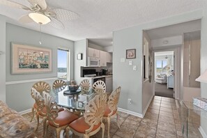A dining area with a glass table and chairs, a ceiling fan, and a kitchen in the background. - Corner Unit w/Sunrise & Sunset Views!| Pool | 1307 (Virginia Beach)