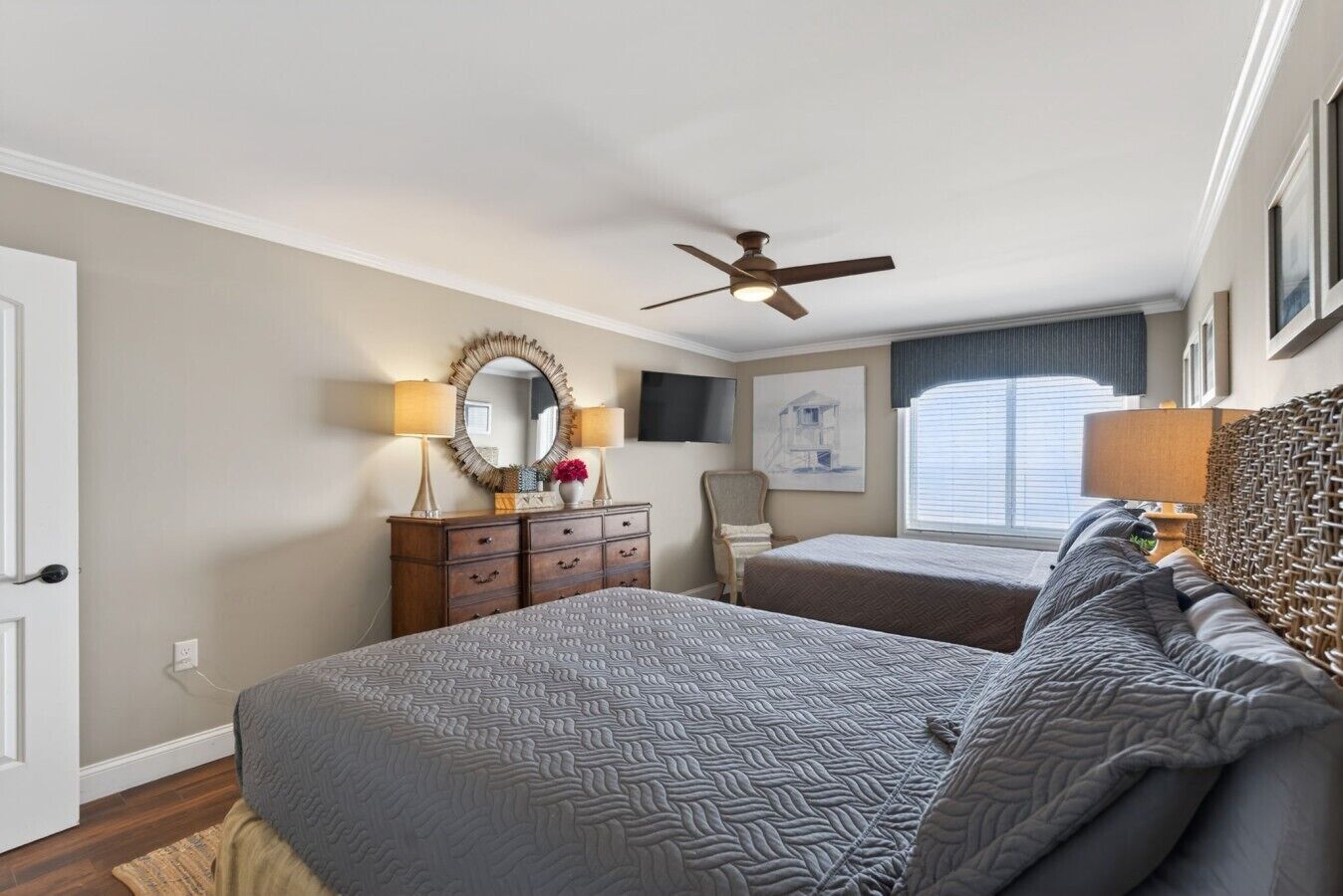 A hotel room with a large bed, a smaller bed, a dresser, a mirror, a ceiling fan, and a window with blinds.
