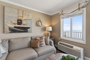 A living room with a beige sofa, a painting of a sailboat, a lamp, and a window with a view of the sea.