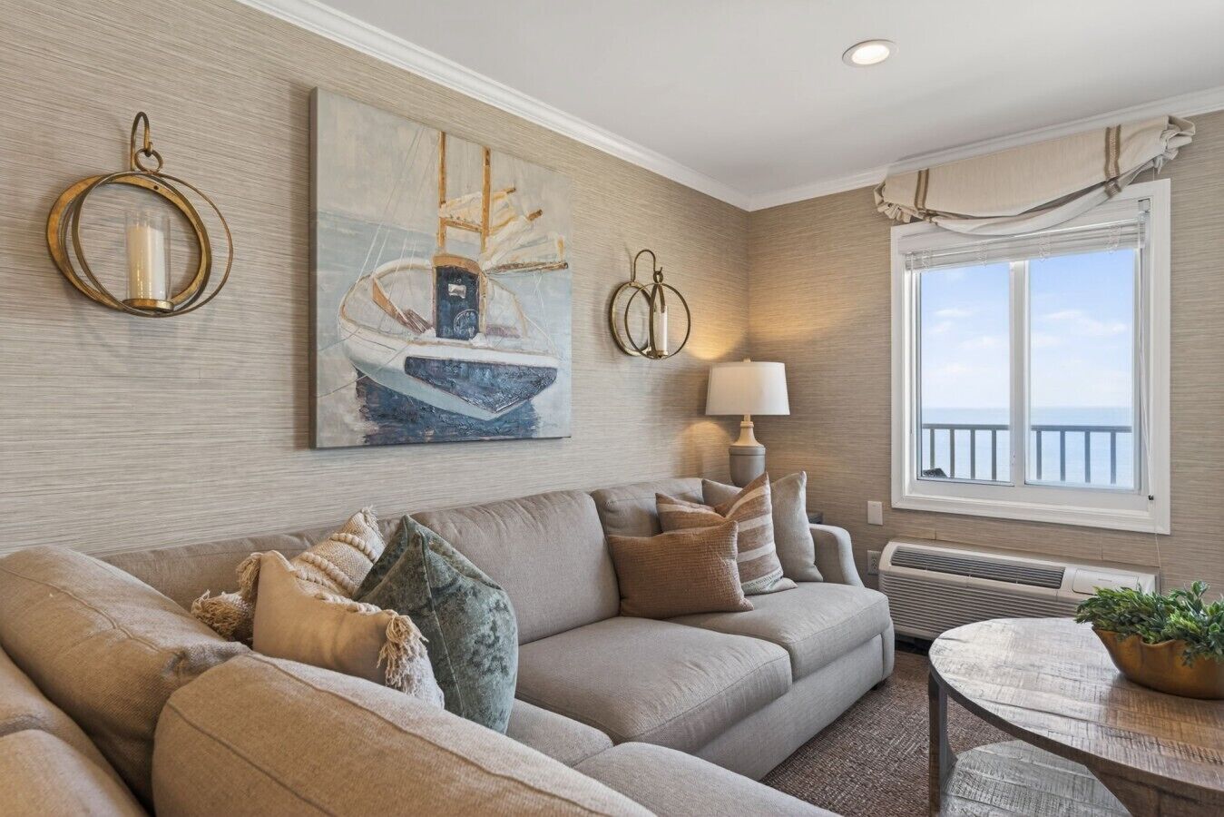 Ett vardagsrum med en beige soffa, en målning som föreställer en segelbåt, en lampa och en utsikt över havet genom ett fönster.