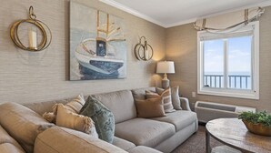 A living room with a beige sofa, a painting of a sailboat, a lamp, and a view of the sea through a window.