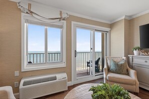 A balcony with a view of the ocean, a beige armchair, a white air conditioning unit, and a flat-screen TV on a cabinet.