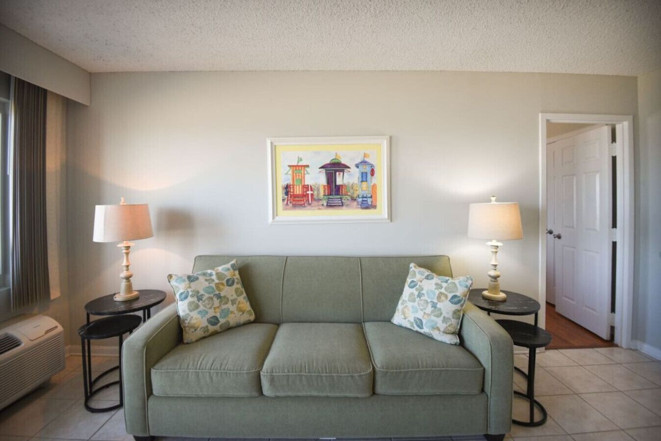 A living room with a green sofa, two side tables with lamps, a framed picture on the wall, and a door in the background.