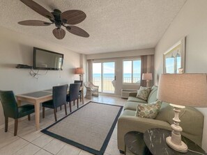 A spacious living room with a dining area, a sofa, a TV mounted on the wall, and a balcony with a view of the beach. - Overlooks Ocean | Dolphin Watching Paradise | Pool (Virginia Beach)