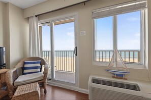 Quarto com vista para a praia, uma cadeira de vime com almofada listrada, um pequeno adorno de veleiro e um ar condicionado branco.
