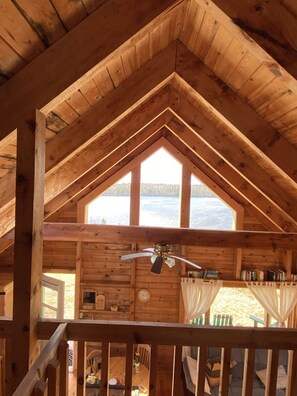 A cabin with a high ceiling, wooden beams, a ceiling fan, a large window with a view of a lake, and a small balcony with a railing.