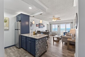 A modern kitchen with a large island, a dining area with a view, and a living room with a sofa and lamp. - Beachwalk Modern Condo Overlooking Ocean Sunshine! (Virginia Beach)