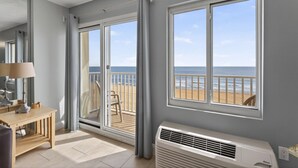 A room with a view of the beach, a sliding glass door, a desk with a lamp, and a white air conditioner.