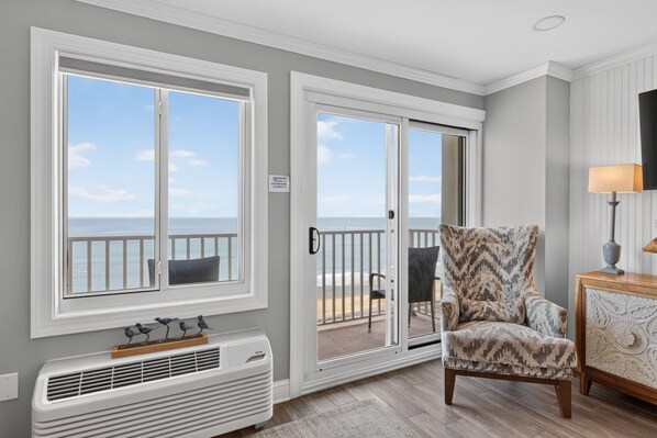 A room with a view of the ocean, a chair, a table with a lamp, and a wall-mounted heater.