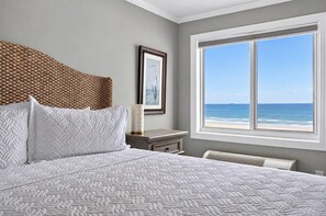 A bedroom with a large bed, a view of the ocean, a framed picture on the wall, and a bedside table with a lamp.