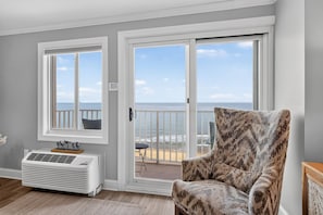A room with a large sliding glass door leading to a balcony with a view of the ocean. There is a white air conditioning unit and a patterned armchair.