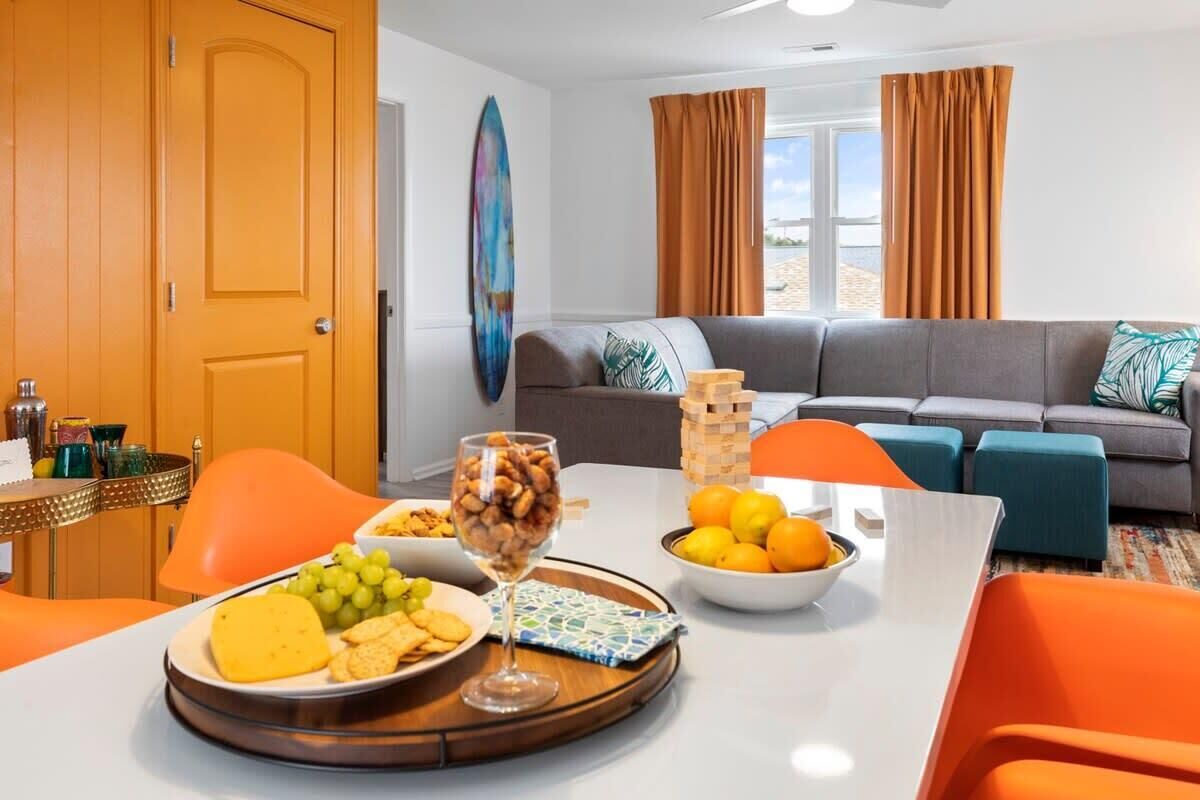 Un salon moderne avec un canapé gris, des fauteuils orange, une table à manger dressée, et une grande fenêtre avec des rideaux.