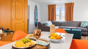 A modern living room with a grey sofa, orange chairs, a dining table with food, and a large window with curtains. - Beach Nest 2 Stylish Condo  2 Min Walk to Beach (North Myrtle Beach)