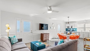 A modern living room with a grey sofa, a coffee table, and a dining area with orange chairs. A kitchen with white cabinets and a microwave is visible in the background. - Beach Nest 2 Stylish Condo  2 Min Walk to Beach (North Myrtle Beach)