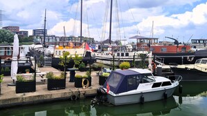 Ein Yachthafen mit verschiedenen Schiffen, ein kleiner Steg mit Kübelpflanzen und eine Stadtkulisse im Hintergrund.