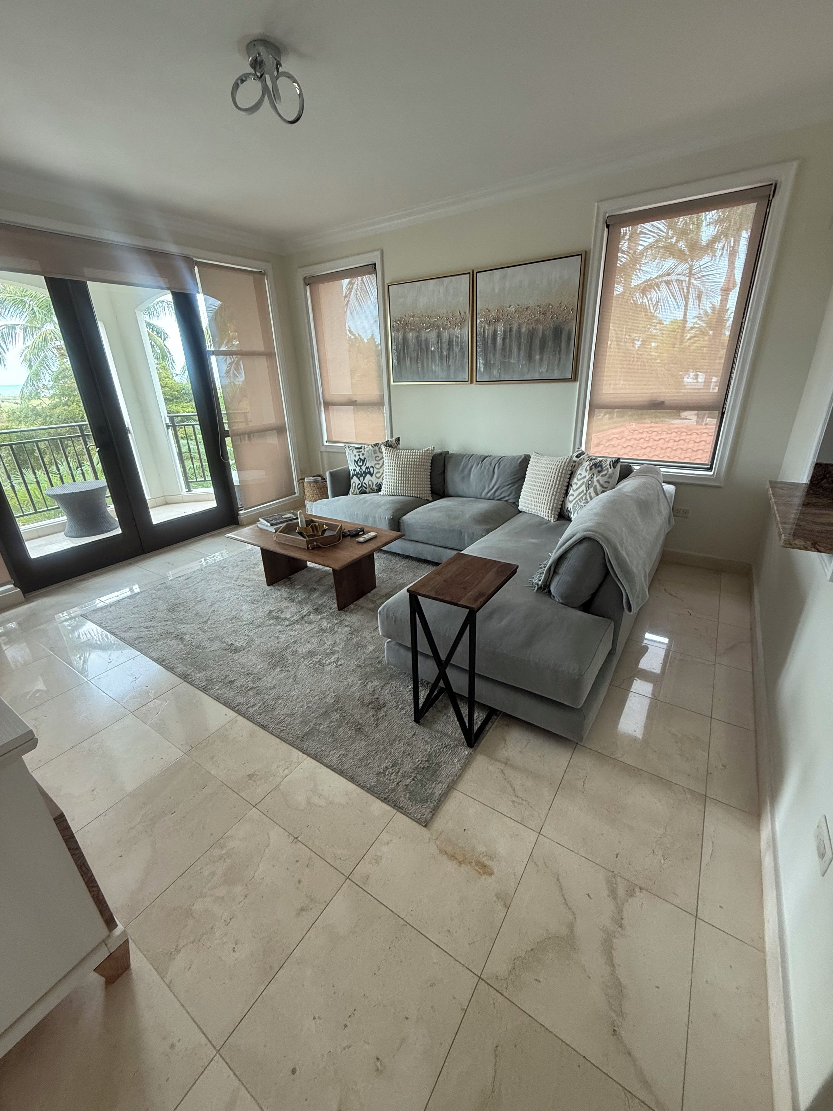 A spacious living room with a grey sofa, a coffee table, and a large window with a view of palm trees.