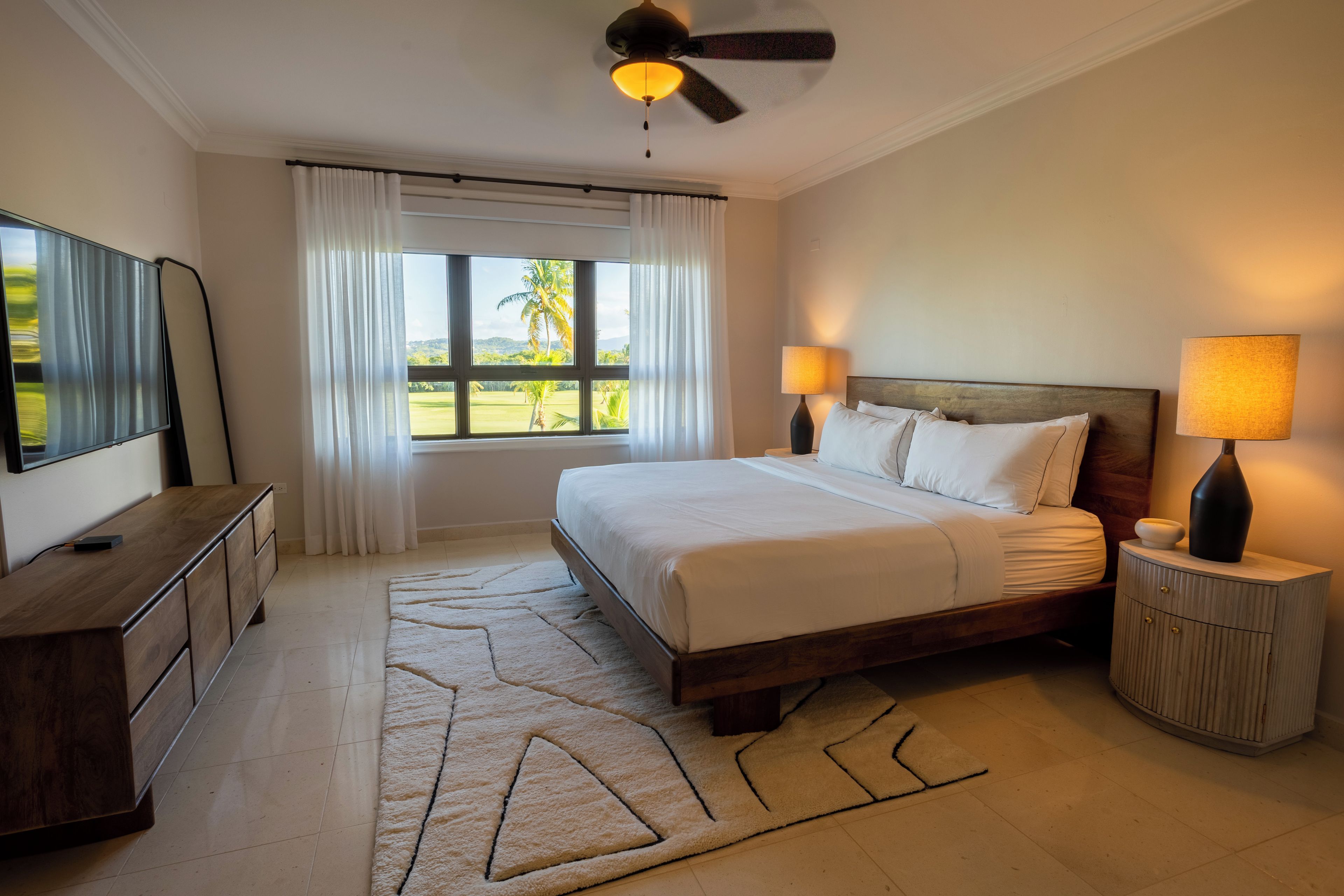 A hotel room with a bed, bedside lamps, a TV, and a view of palm trees.