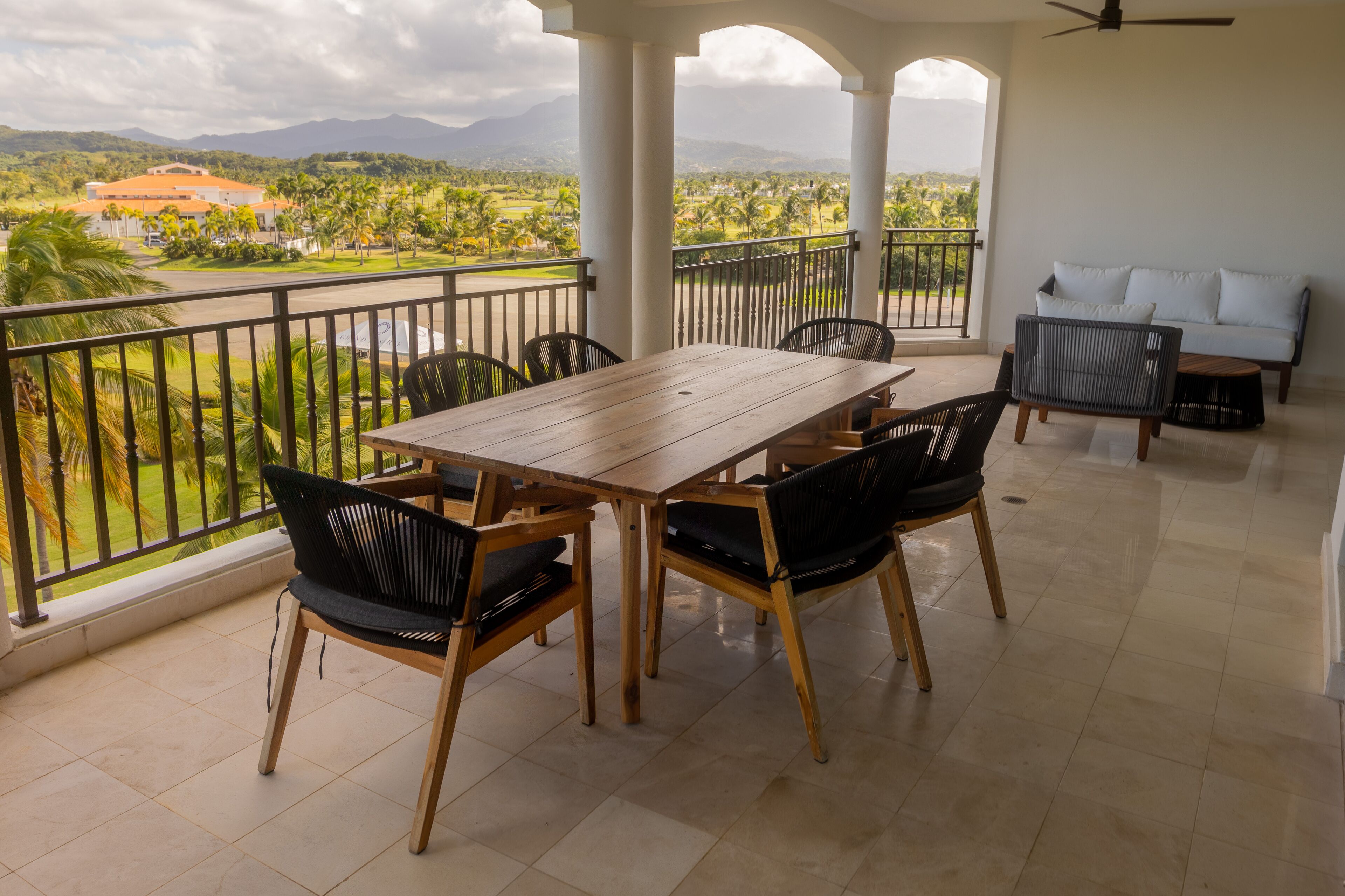 A balcony with a wooden table and chairs, a sofa, and a view of a green landscape with mountains in the distance.