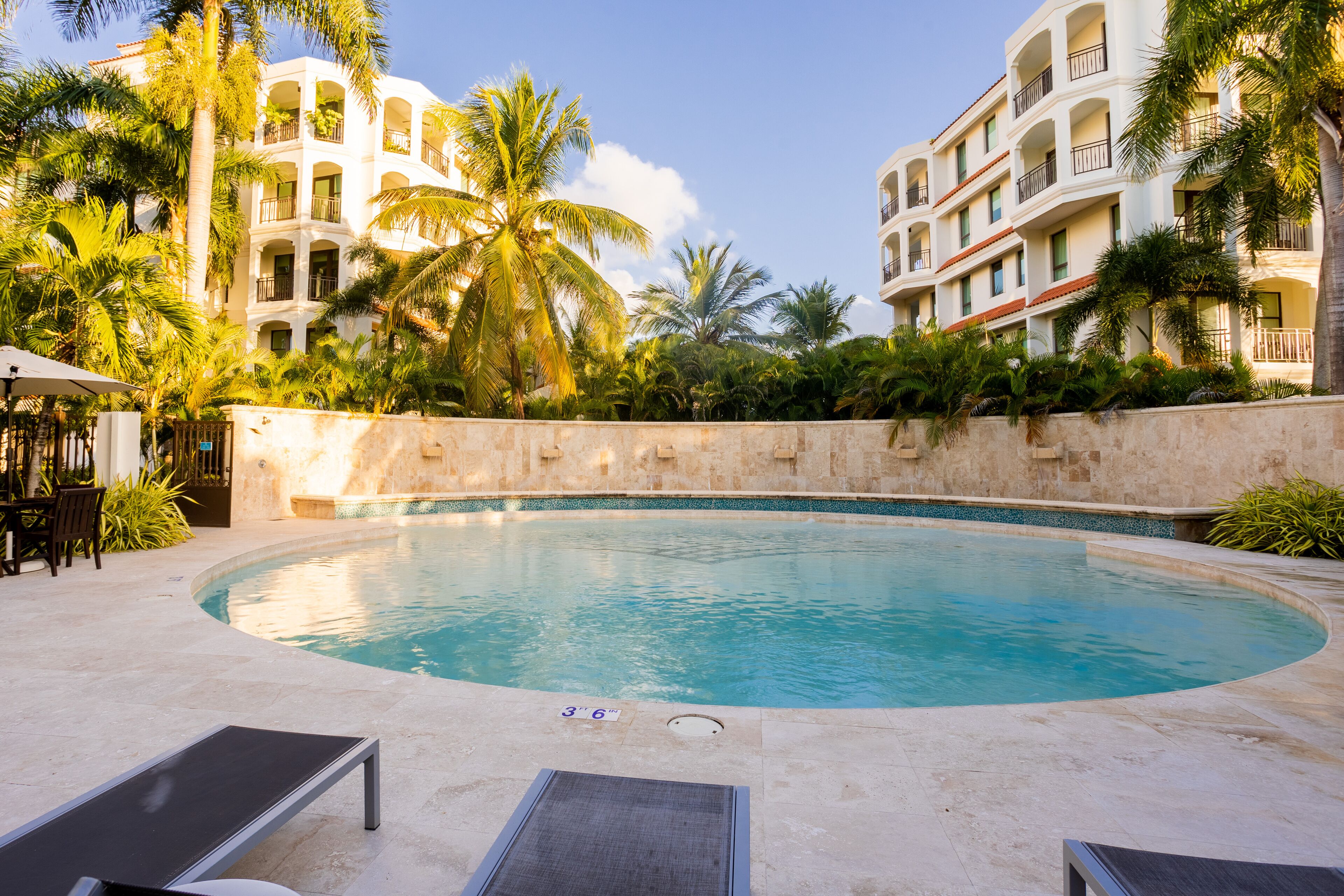 Una piscina con sillones de descanso, rodeada de palmeras y un edificio con balcones.